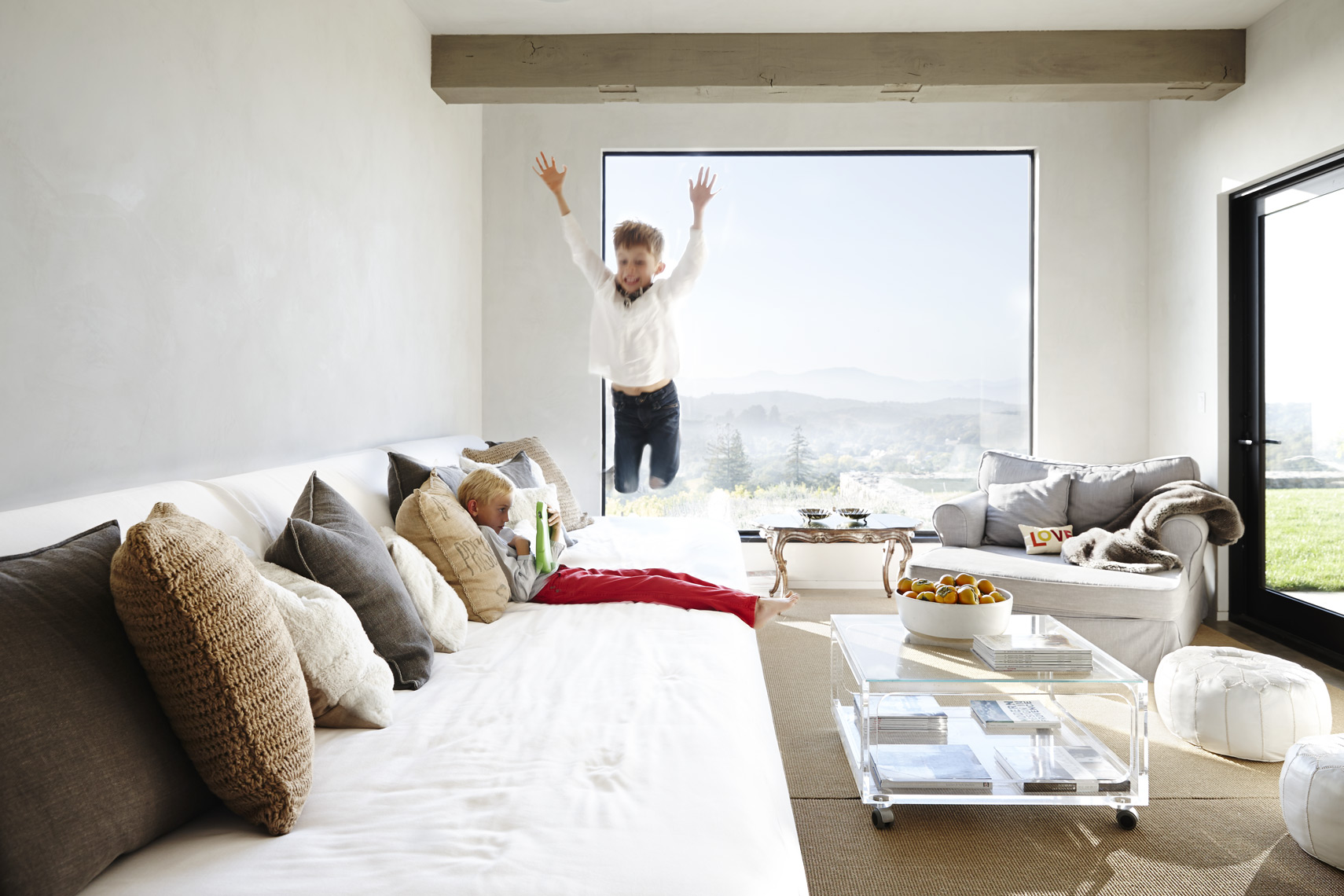 Child leaping over kid using ipad on sectional sofa in modern house Sean Dagen Photography